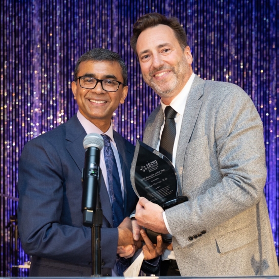 short man with glasses stands at podium holding trophy with taller man with goatee in tan suit