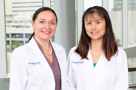 Two women in lab coats standing against a windowed backdrop