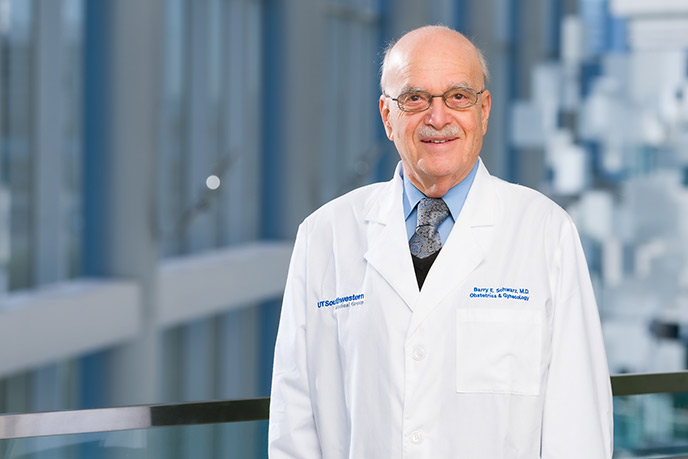 Bald man with mustache, glasses, wearing a labcoat in a glass-lined hallway