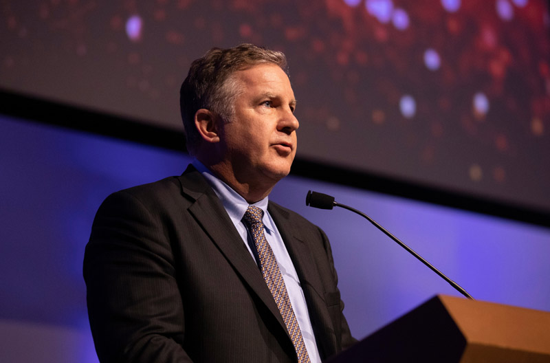 Man standing at lectern speaking into microphone, wearing a suit