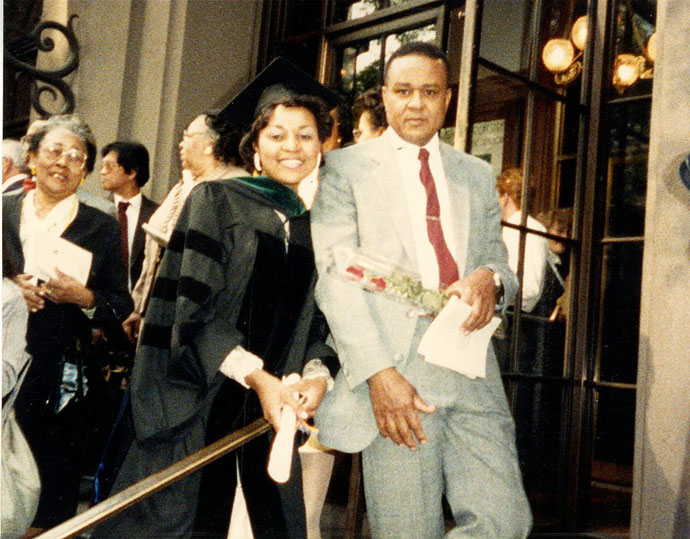 Woman wearing black graduation cap and gown standing next to a man with a red tie and suit on