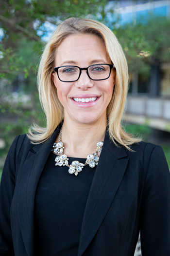 Woman standing in front of a tree, she has blonde hair, wearing glasses, and a black blazer