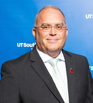 Man short white hair, glasses, wearing a dark suit