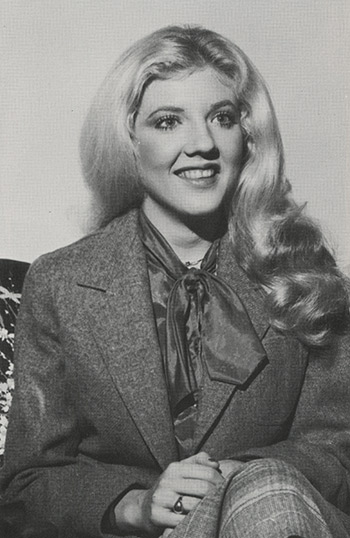 Black and white photo of a woman with long blonde hair, silk scarf, and textured blazer
