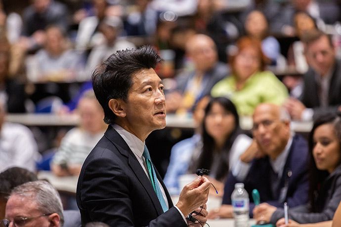 Man with black hair, wearing a suit, speaking into a small microphone