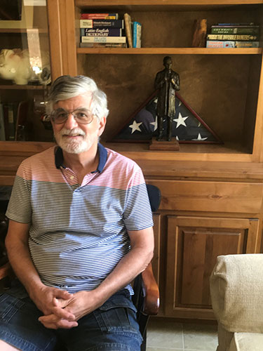 Dr. Paul Vanatta sits in a chair given to him by the late Dr. Ver¬nie Stembridge, former UTSW Chair of Pathology. In the back¬ground is an American flag that covered his father’s casket at his funeral. The statue, which he inherited from his father, is called “The Mentor” and was sculpted by a fellow UT South¬western Medical School graduate.