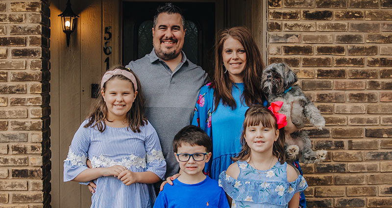 Family of two parents and three children standing in front of house smiling