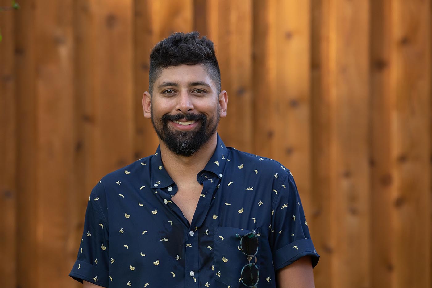 Man with dark hair, beard in blue buttoned shirt smiling