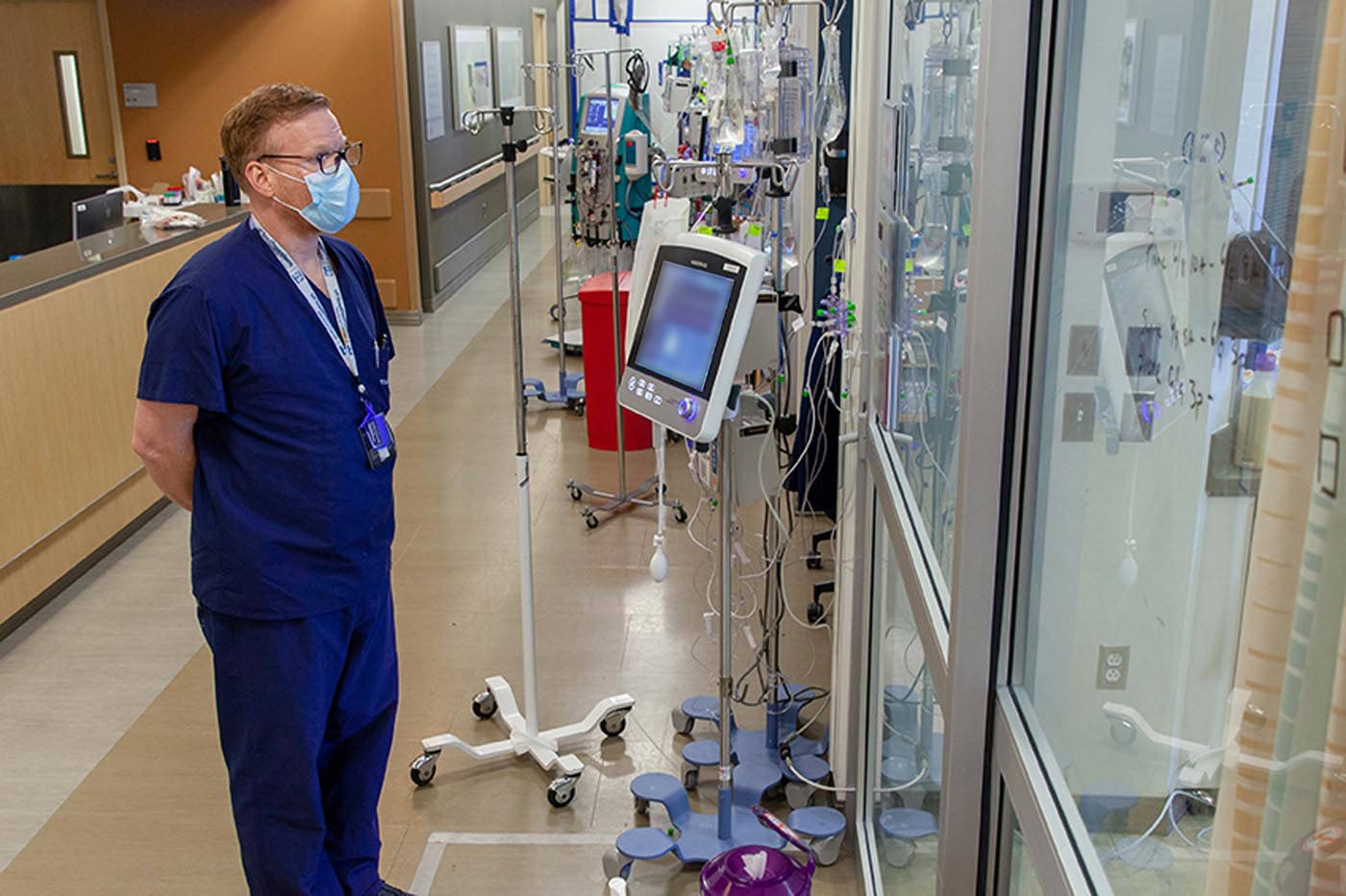 Man in scrubs and mask looking through glass wall, surrounded by medical equipment