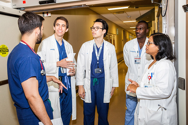 Four people in white lab coats and one in blue scrubs talking in a hallway
