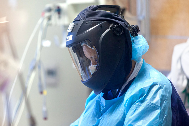 Person in scrubs, gown, mask, and black helmet and mask caring for a patient