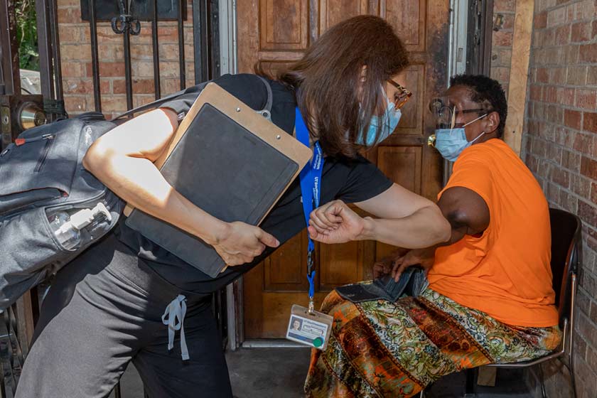 Woman in mask giving elbow bump to another masked woman sitting on a porch