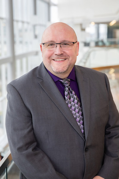 Man with glasses, wearing grey sports jacket and purple tie