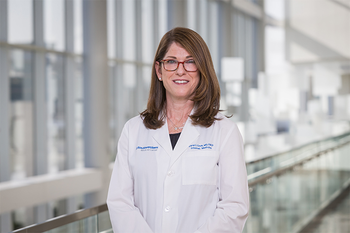 Woman with dark hair, glasses, wearing a labcoat standing in a window-lined hallway