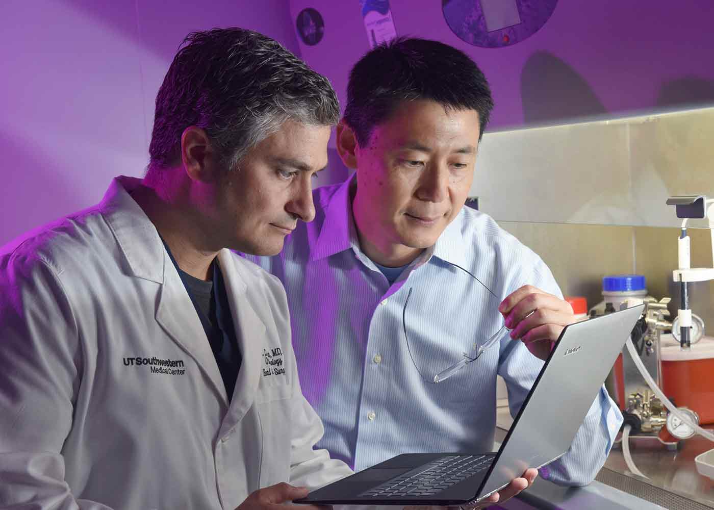 Two men, one in white lab coat and the other in light blue button down, looking at laptop screen.