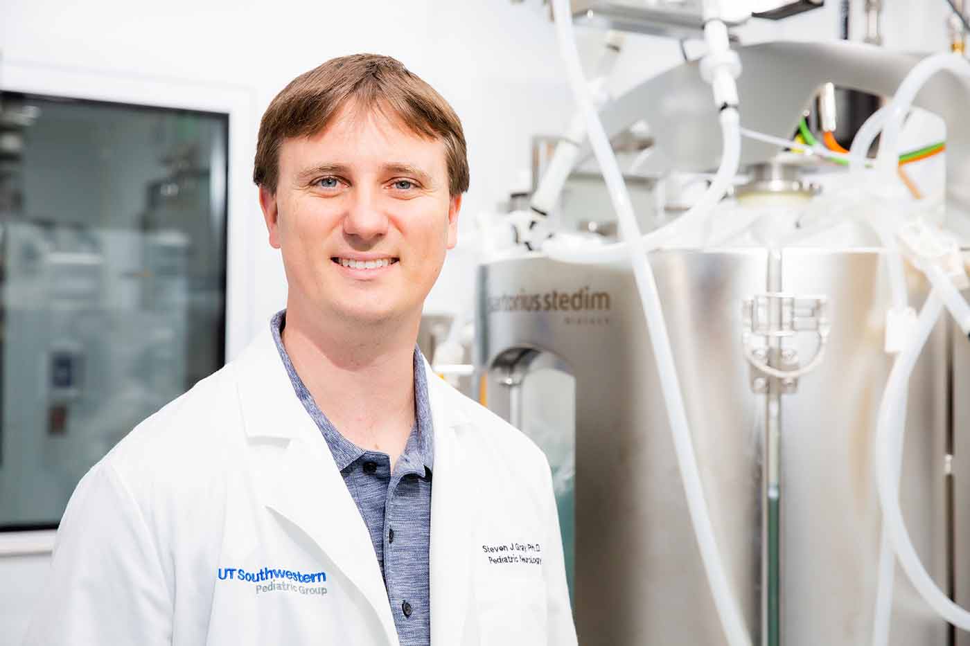 Man in blue shirt, white lab coat, standing in front of metal tank