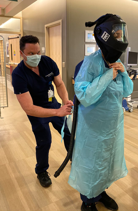 Man in scrubs tying gown on woman wearing PPE helmet