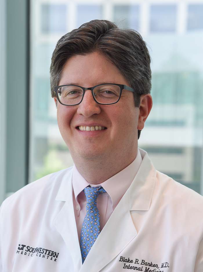 Man with dark hair parted hair, glasses, wearing a lab coat and blue tie