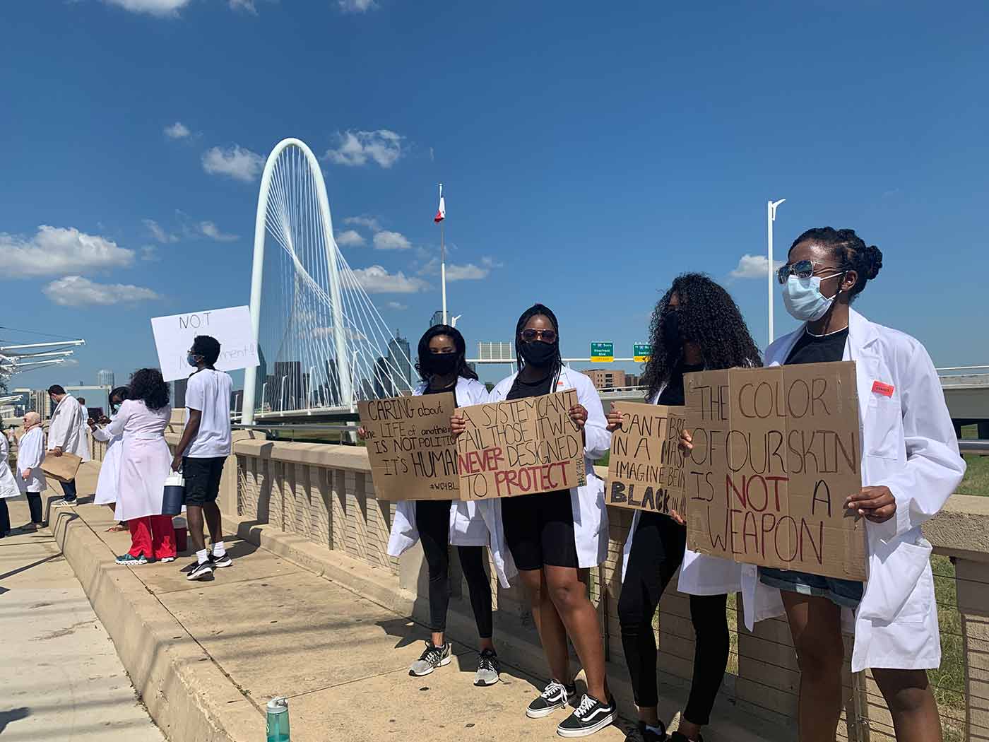 Black women in white lab coats holding protest signs on bridge.