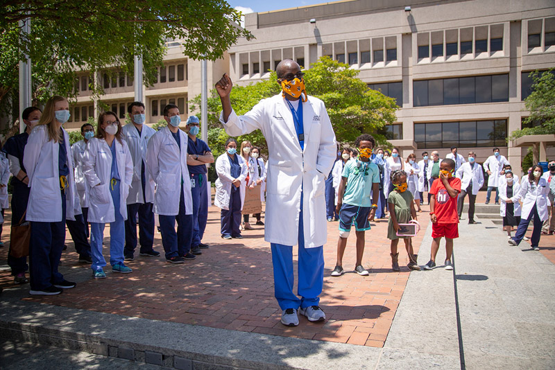 Black man in scrubs, white lab coat, wearing orange mask, speaking to crowd