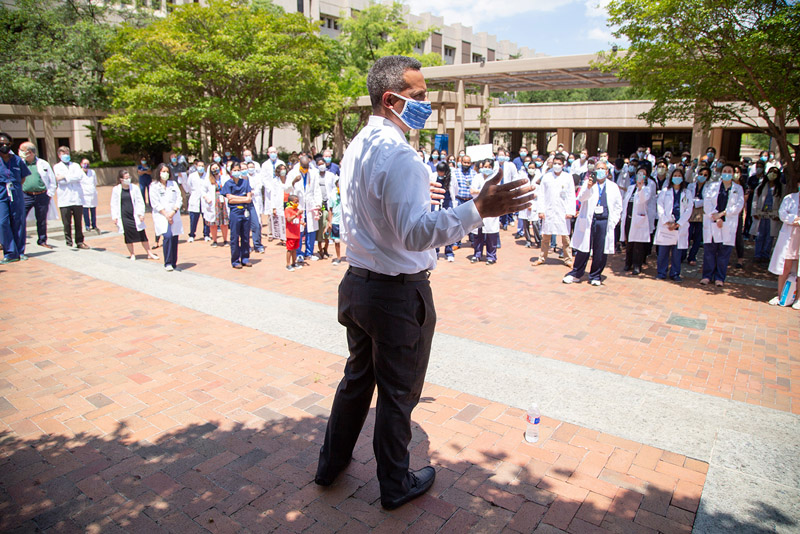 Black man with mask, in dress shirt and slacks speaks to crowd