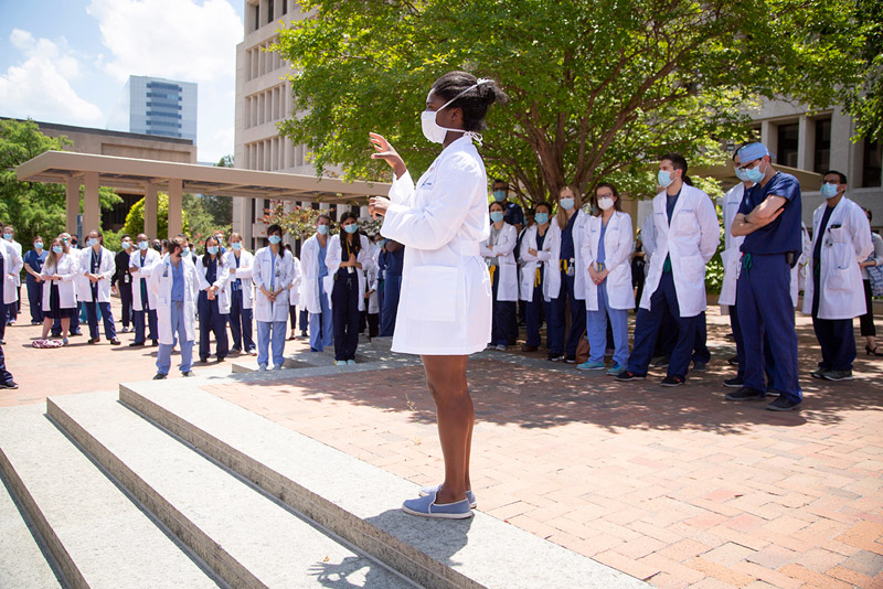 Black woman in mask, white coat, speaking to crowd