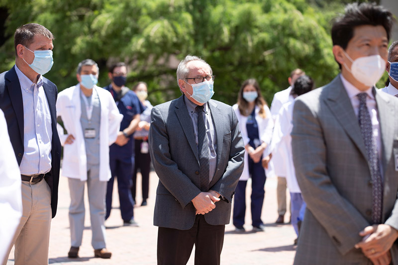 People in suits and masks standing and listening amongst crowd of people