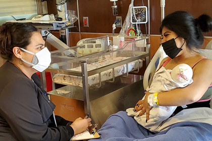 two masked women, one holding baby, sit in nursery