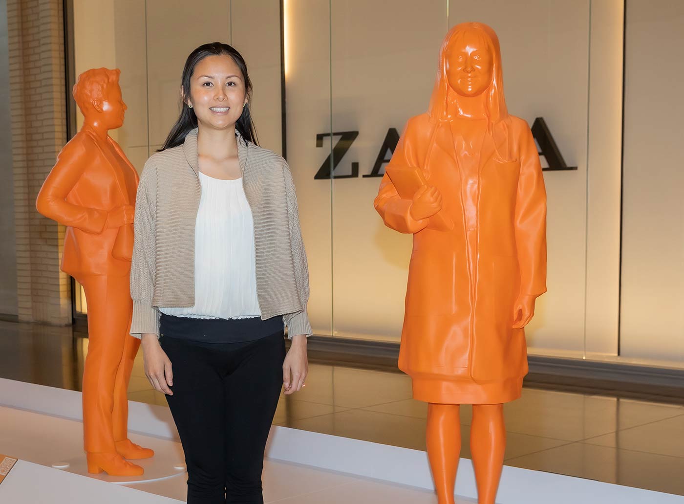 Woman in tan jacket, white shirt and black pants standing by statue of herself