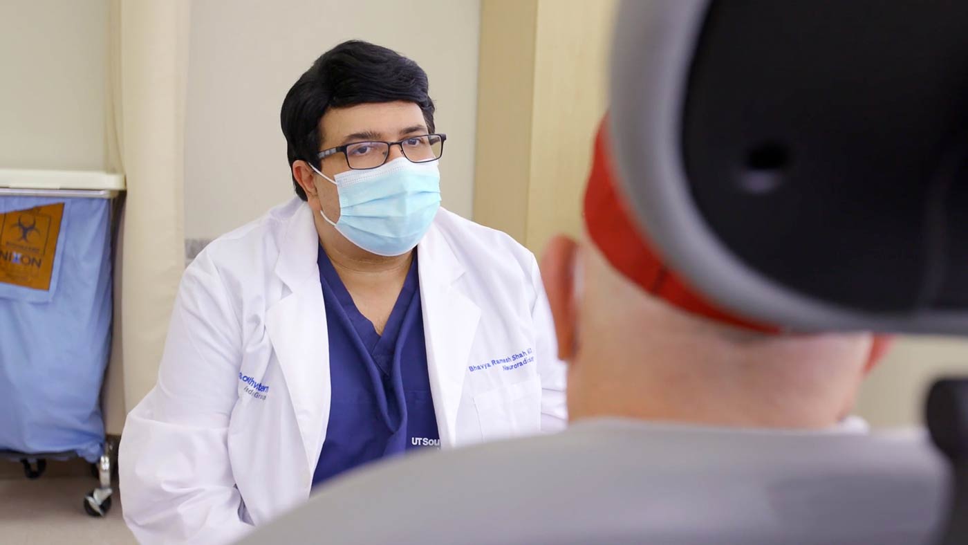 Man with glasses, whtie lab coat, and mask talking with a person