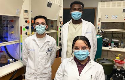 three students wearing white coats in lab