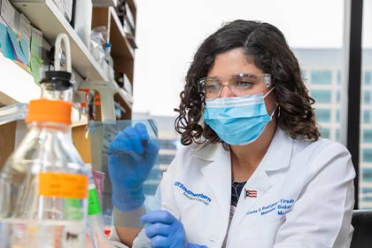 Dark-haired woman wearing a lab coat, paper mask, and blue latex gloves working in lab