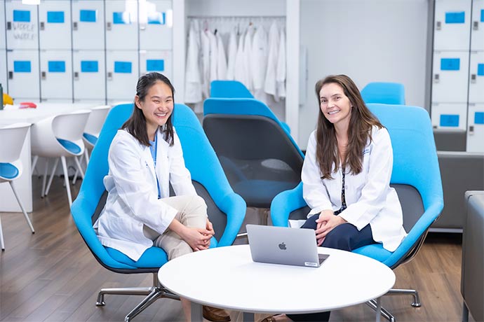 two women in white lab coats sitting in blue chairs