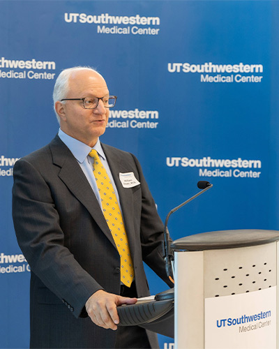 man in dark suit, blue shirt, yellow tie at podium 