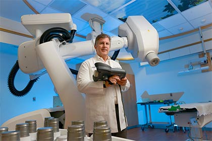 Man wearing lab coat holding a small piece of equipment, standing in front of a large medical machine