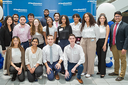 diverse group of 17 undergraduate students pose in front of UTSW backdrop and blue and white balloons