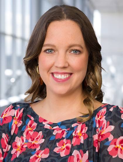 woman with grey, black, red, pink, and blue blouse, shoulder-length dark brown hair with highlights