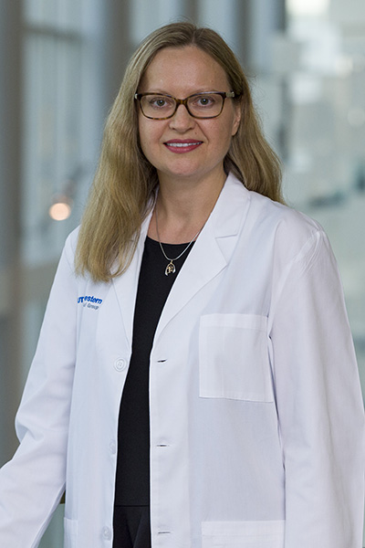Smiling woman with long blond hair and dark-rimmed glasses, wearing a lab coat over a black blouse and slacks.