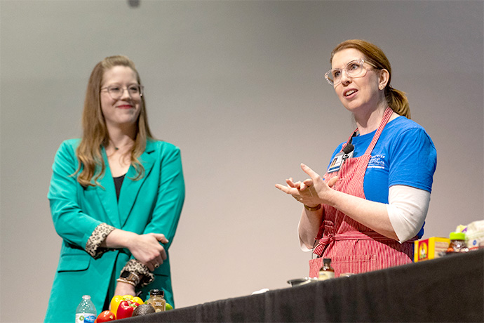 Two smiling women standing behind a table full of healthy snacks, including fruit.