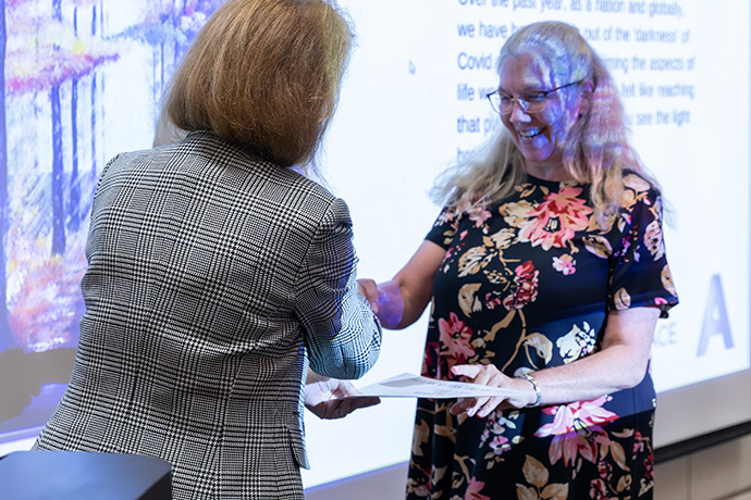 Smiling woman with fair hair and glasses shaking hands with woman with red hair wearing a checked jacket. A picture of a forest is projected on the wall and partially on the fair haired woman's head.