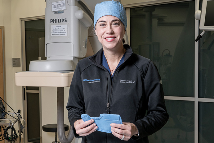 Dr. Kirkwood, smiling woman wearing a UT Southwestern Medical Center jacket and blue surgical cap, with medical equipment in the background.