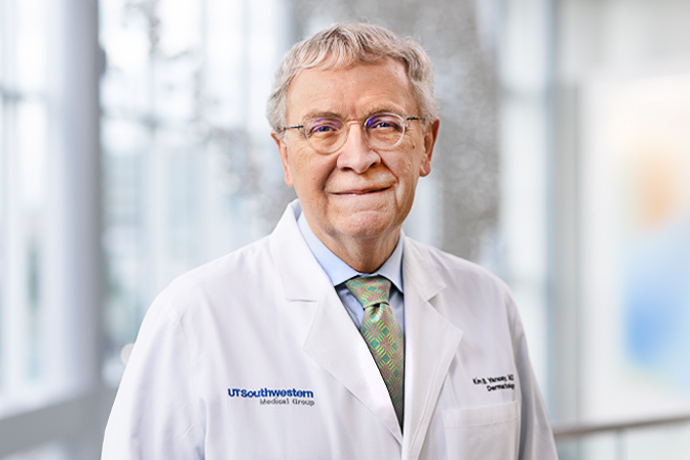 headshot of elderly male doctor with grey hair and glasses wearing white lab coat over blue shirt and patterned tie