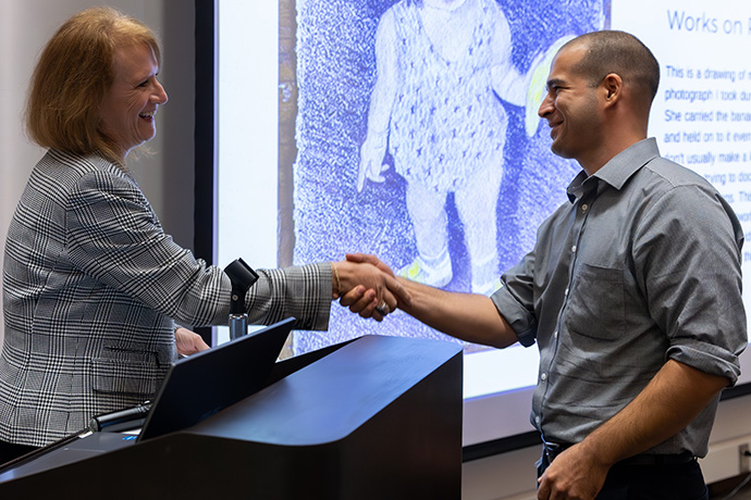 Woman with red hair and checked jacket shaking hands with a man with closely cropped short hair wearing a blue shirt standing in front of a blue sketch of a small child holding a banana.