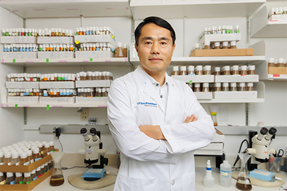 Dr. Pan, a man with dark hair wearing a lab coat, standing in front of shelves in the lab.