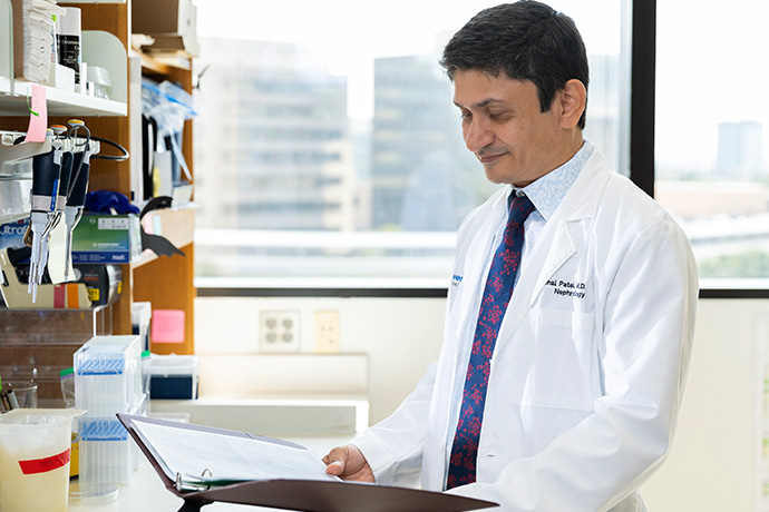 Dr. Patel, man with dark hair wearing a lab coat standing in a lab looking down at a notebook.