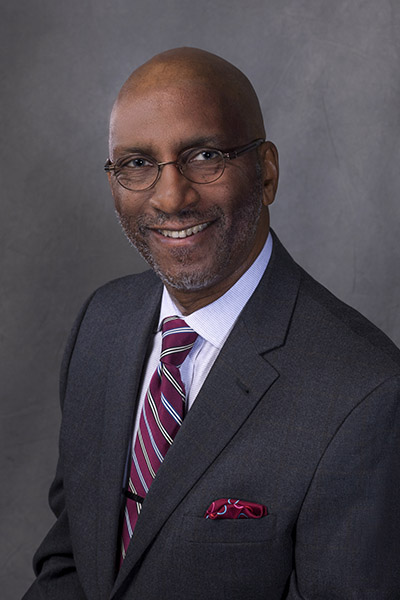 Smiling man with shaved head and trim beard and mustache, wearing a dark gray suit, white shirt and red striped tie.