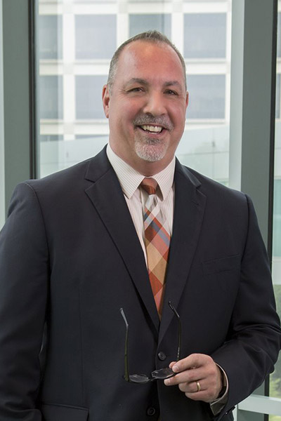 Smiling man with light hair and gray beard and mustache, wearing a dark gray suit, white shirt and plaid tie.