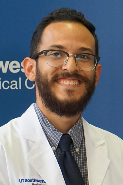 Smiling man with dark hair, beard and mustache, wearing a lab coat and glasses.
