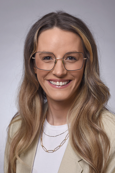 Smiling woman with long brown hair, wearing a tan jacket and glasses.