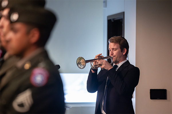 man in suit play bugle with military color guard standing nearby